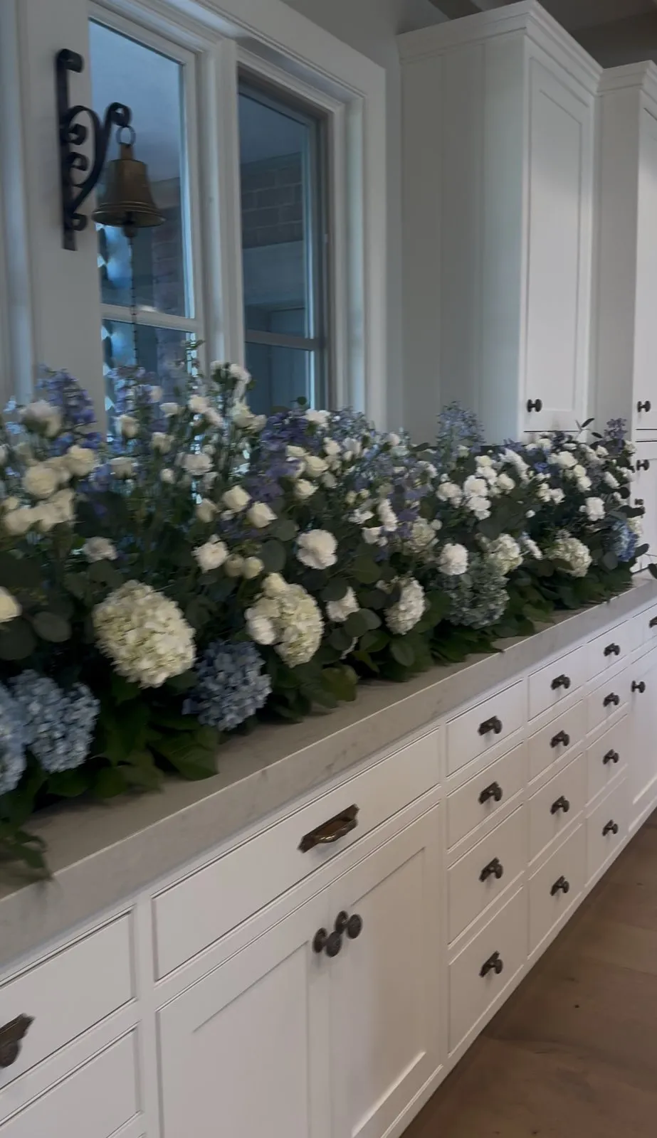 A long, lush floral arrangement of blue hydrangeas, white roses, and tall blue delphiniums on a kitchen counter.