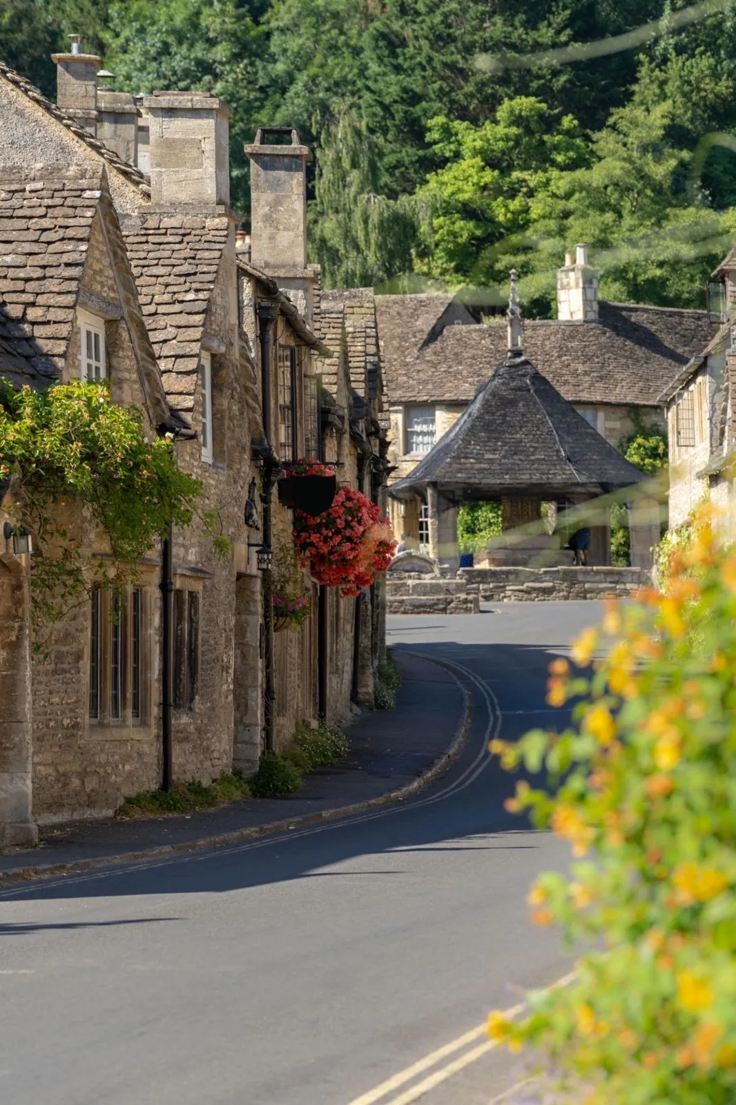 Narrow road winding through a historic Cotswolds village lined with stone houses and flowers.