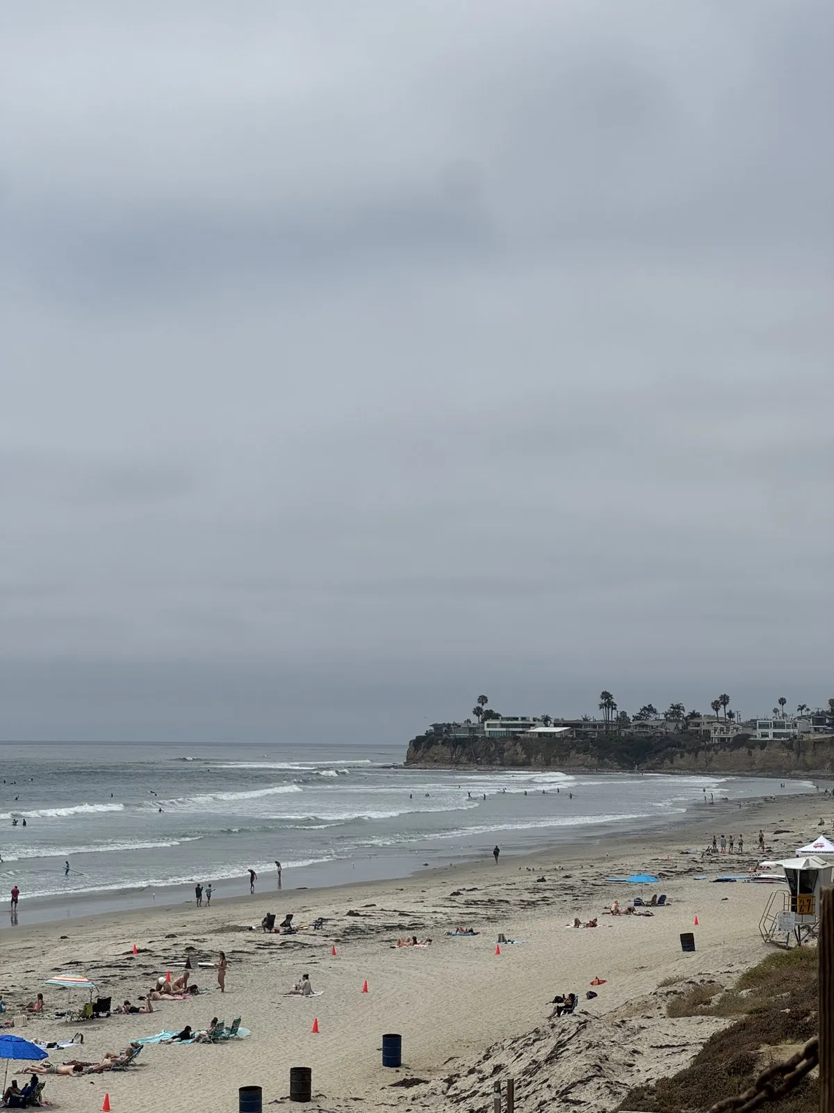 An overcast coastal beach view with sand, ocean waves, and houses along a bluff.